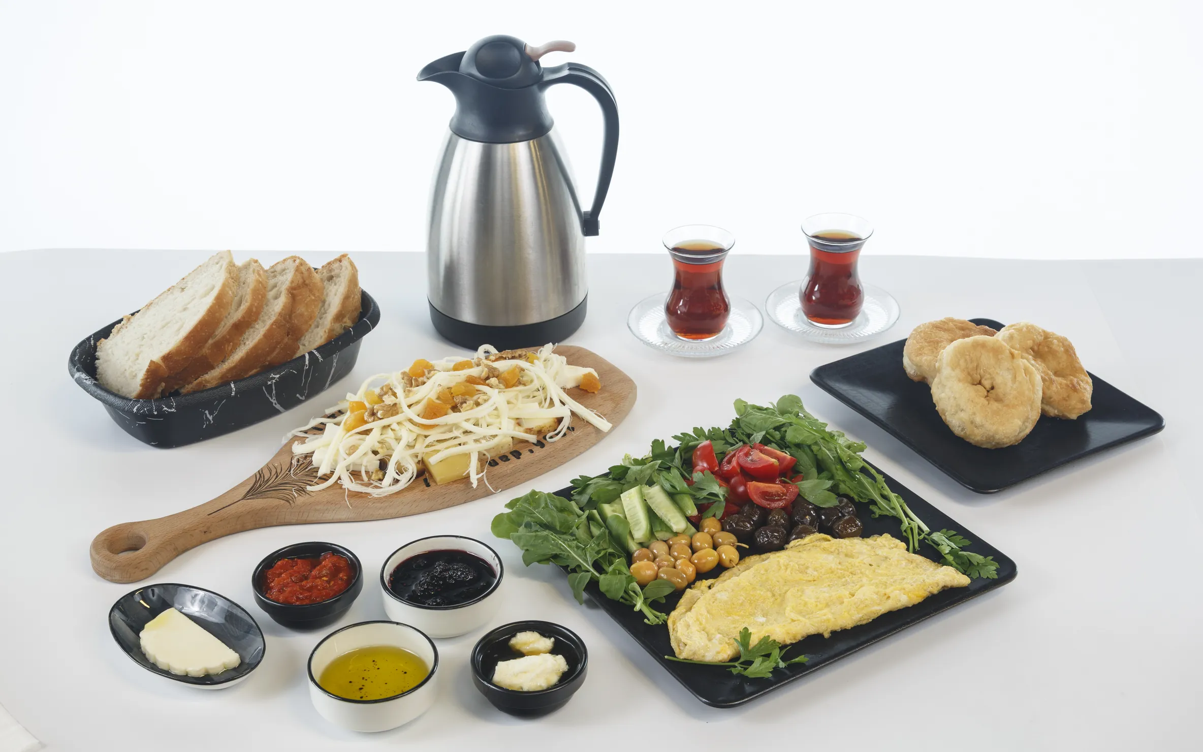 İki kişilik serpme kahvaltı sofrası: çelik termos, iki bardak çay, ahşap tahtada ceviz ve kayısılı tel peynir, siyah tabakta omlet, roka, maydanoz, salatalık, kiraz domates ve iki çeşit zeytin; yanında ekmek sepeti, pişi, tereyağı, iki reçel, zeytinyağı ve bal.