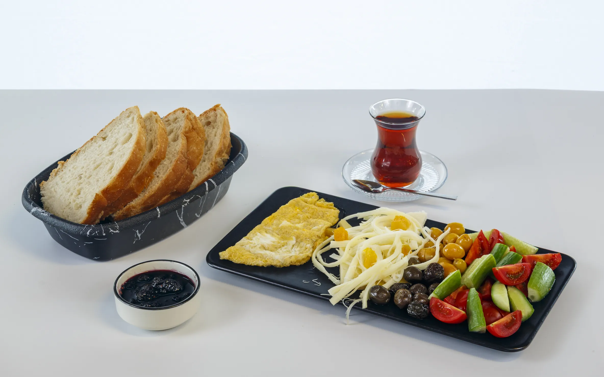 Tek kişilik kahvaltı tabağı: siyah dikdörtgen tabakta omlet, kayısılı tel peynir, yeşil ve siyah zeytin, domates ve salatalık dilimleri; yanında ekmek sepeti, bir bardak çay ve böğürtlen reçeli.