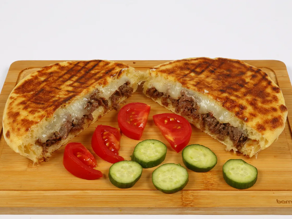 Bambu sunum tahtasında ikiye bölünmüş kavurmalı tost: ızgara izleri belirgin, kesit yüzeyinde kavurma ve eriyip uzayan kaşar görünüyor; ortada domates dilimleri ve salatalık halkaları.