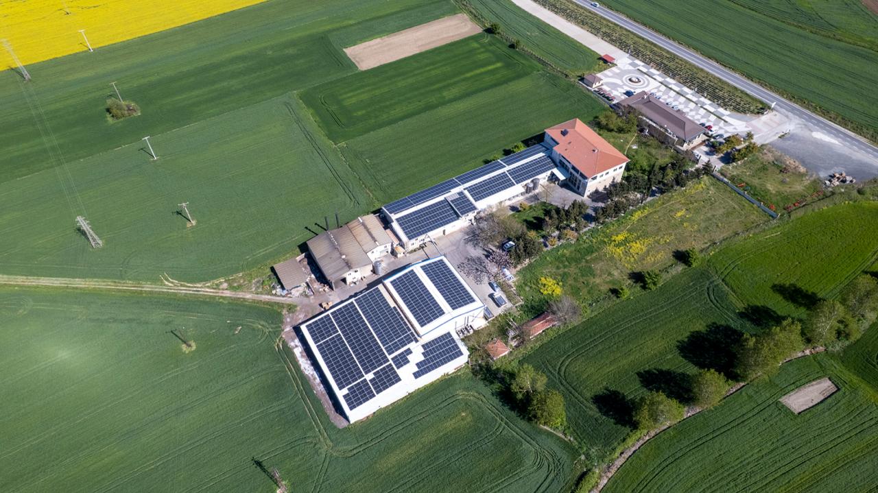 Vize'deki Mandıra Mola tesisinin dron çekimi: çatıları güneş panelleriyle kaplı üretim binaları, yanında satış ve kahvaltı salonu ile otopark, çevrede yeşil ekin ve sarı kanola tarlaları.